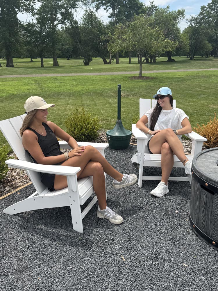 2 women chat while sitting outside on white chairs